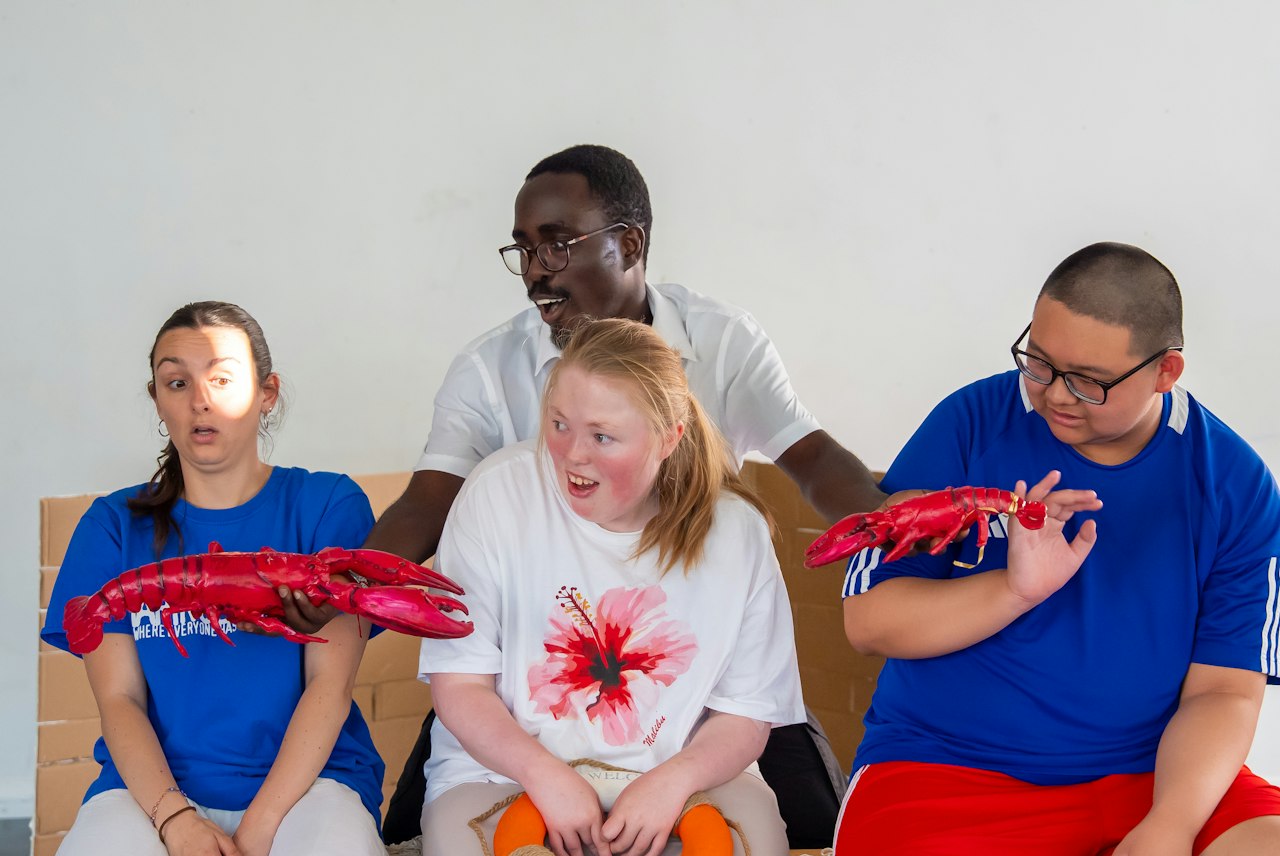 A photo of four young adult performers from Haringey Shed against a white background, wearing expressions of surprise. Two wear white t-shirts, two wear blue t-shirts, and two hold hot pink lobsters.