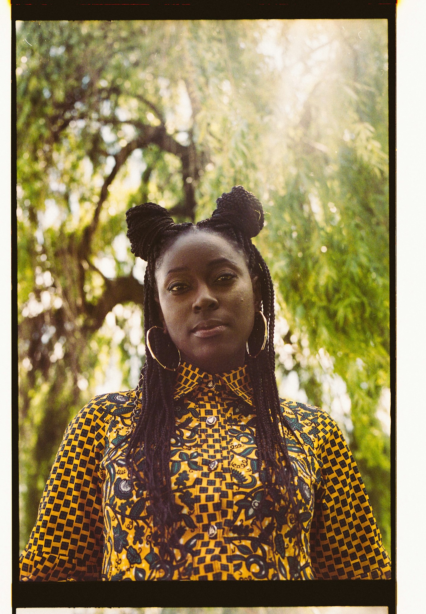 A portrait-style photograph of a young Black woman. She wears a colourful yellow and black printed shirt and has her hair in two braided buns ion top of her head. She is stood outside on a sunny day.
