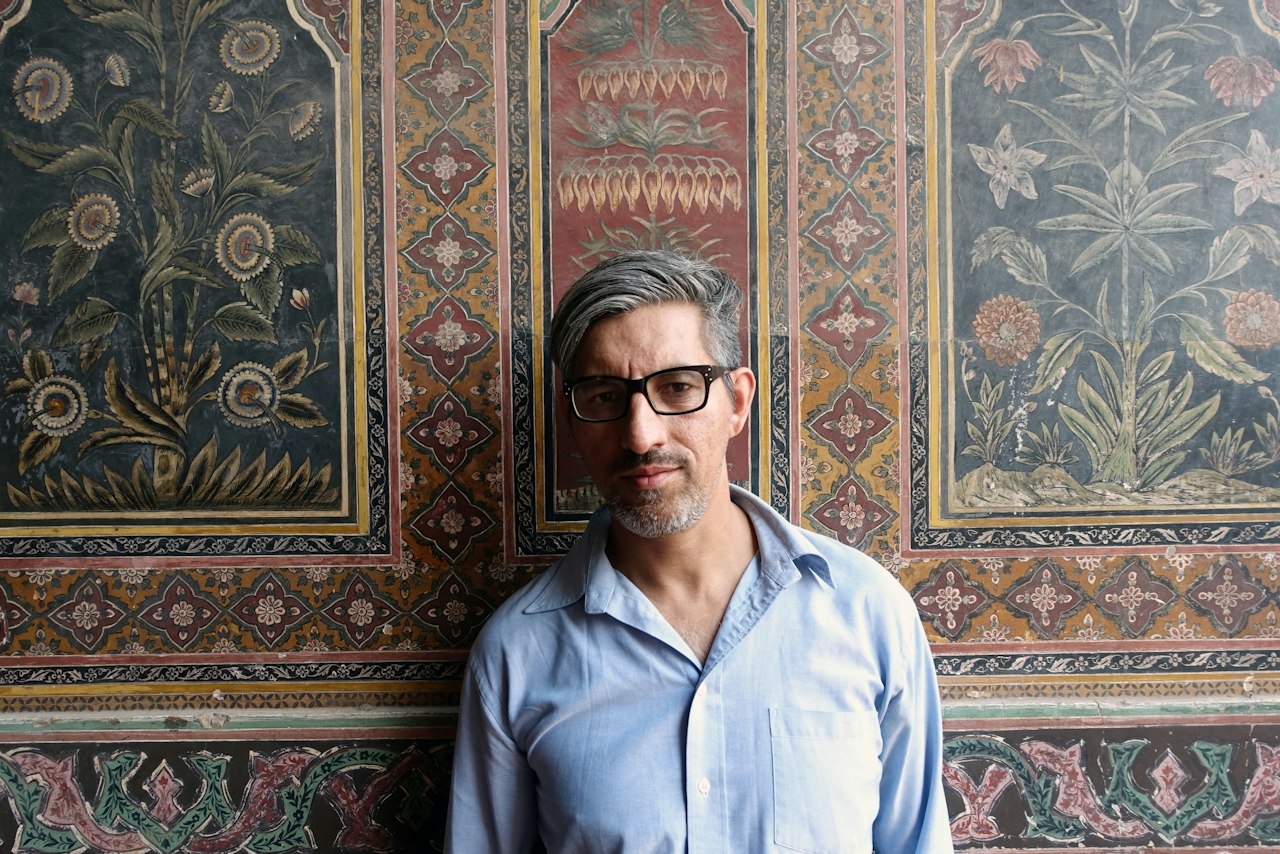 A photograph of the artist Wajid Yaseen. He has as stylish haircut, thick rimmed glasses, and wears a blue linen shirt slightly undone. He is standing against an intricately woven tapestry in shades of orange, red, green and blue.