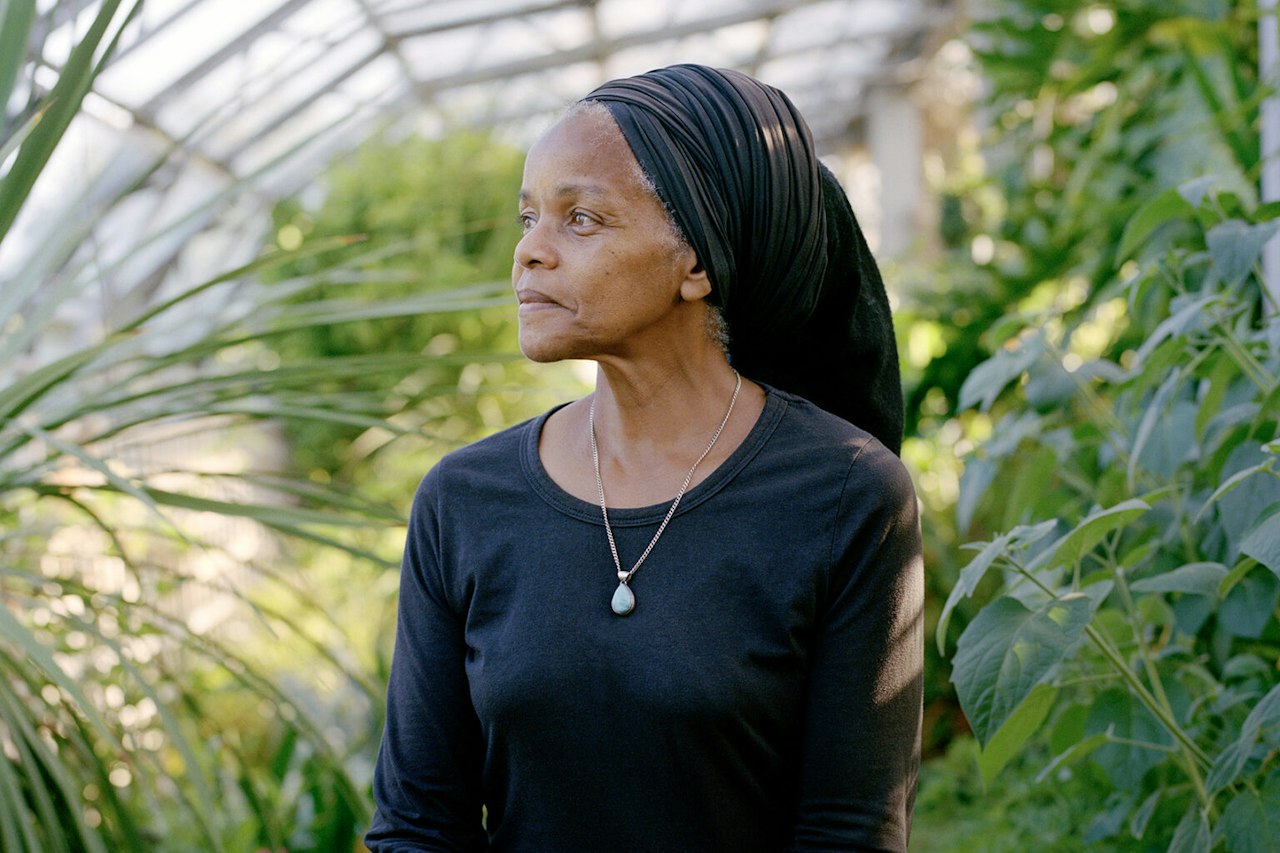 A portrait-style photograph of the artist, Paulette Henry. She is sat outside in a greenhouse - the background is warm and full of palm leaves. Her hair is tied in a black wrap which drops down elegantly  over her shoulders, and compliments her black t-shirt. She turns her face to one side, smiling serenely as she looks into the distance off-camera.