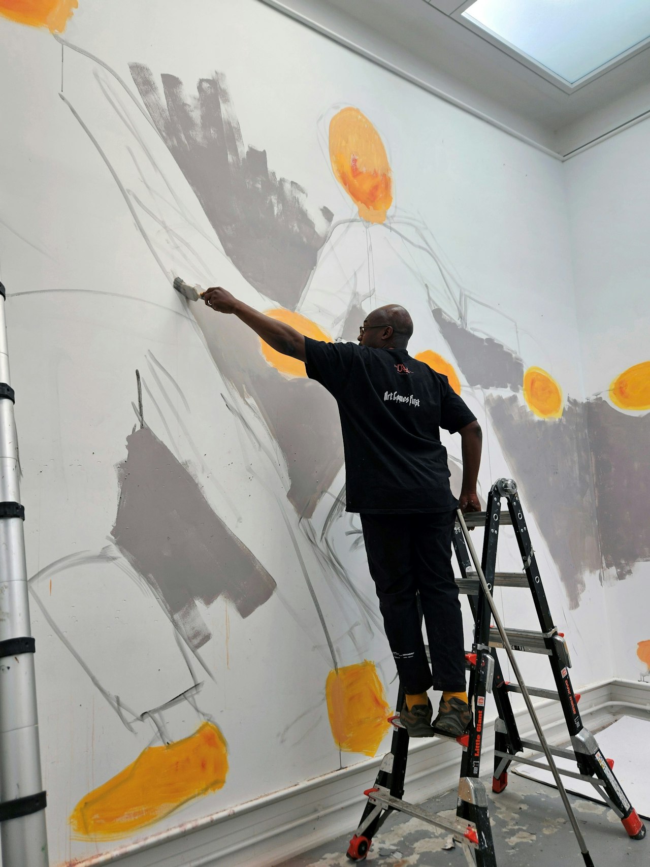 A photograph of a man in a black sweater and trousers. he is stood on a ladder, using a large paintbrush to paint a colourful mural on a wall.