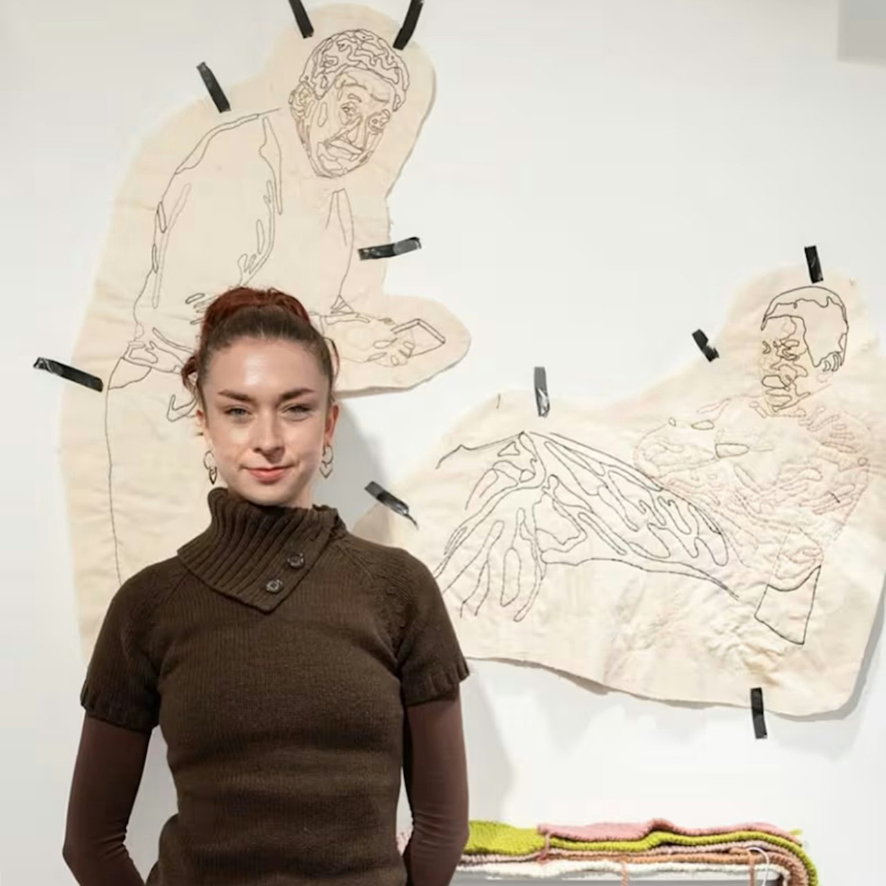 A young woman in a brown jumper with her hair in a ponytail. She stands in front of a large scale artwork of two pencil drawings which are taped up onto a wall.