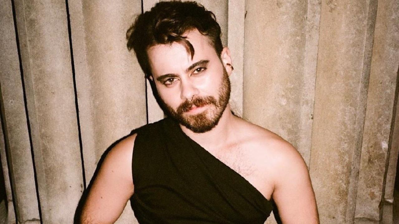 A photograph of a young non-binary person wth long, dark curly hair and a beard. They are wearing dramatic dark eyeliner and a one shoulder black evening top.