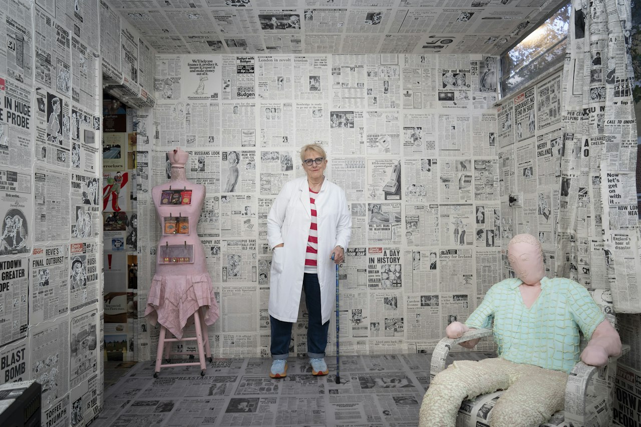 A photograph of the artist Bobby Baker. They are wearing a long white duster coat over a striped red t-shirt and black trousers, and stand with the assistance of a cane. they are stood inside an immersive installation of their own design, which looks like many different news articles from a paper pasted to an interior to look like wallpaper. two mannequins are present either side of the artist.