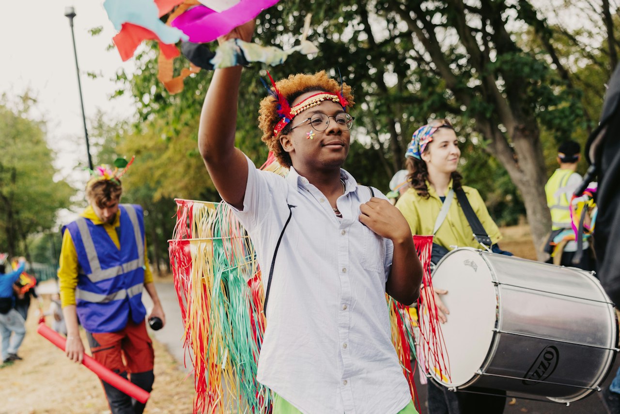 A parade of brightly-dressed people at the Tottenham Carnival Fiesta, walking through a park. The figure at the front waves a streamer in the air, whilst someone else carries a drum.