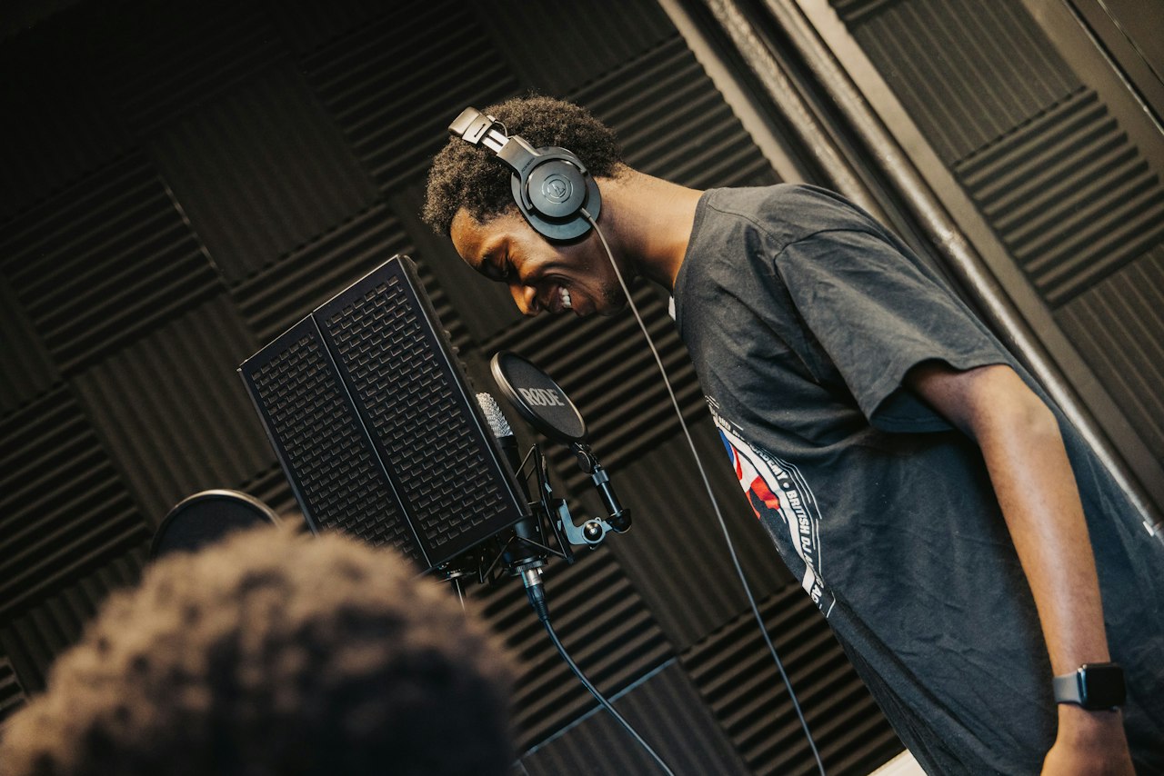In a recording studio, a person stands in-front of a microphone. They have their eyes closed, beaming with a big grin.