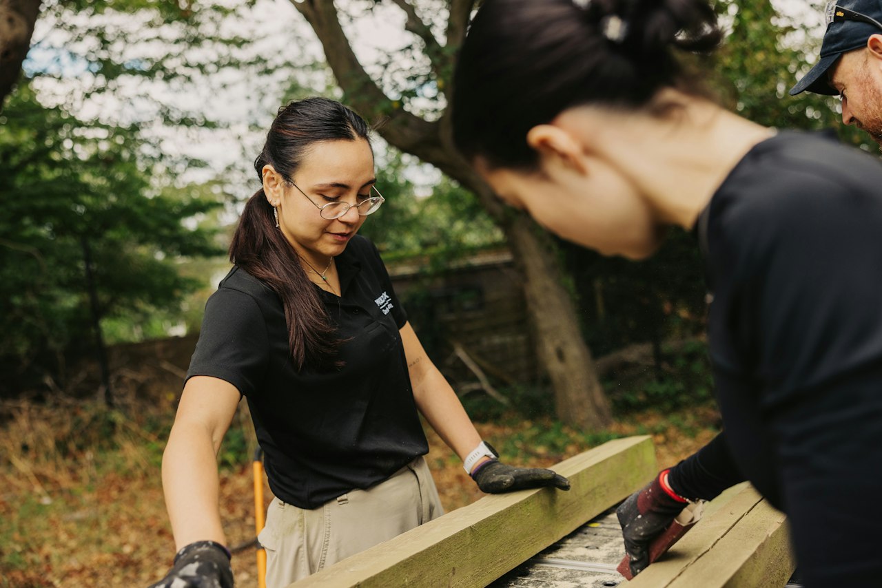 a person in a polo shirt and sensible cargo trousers instructs another person how to align a piece of wood for carpentry.