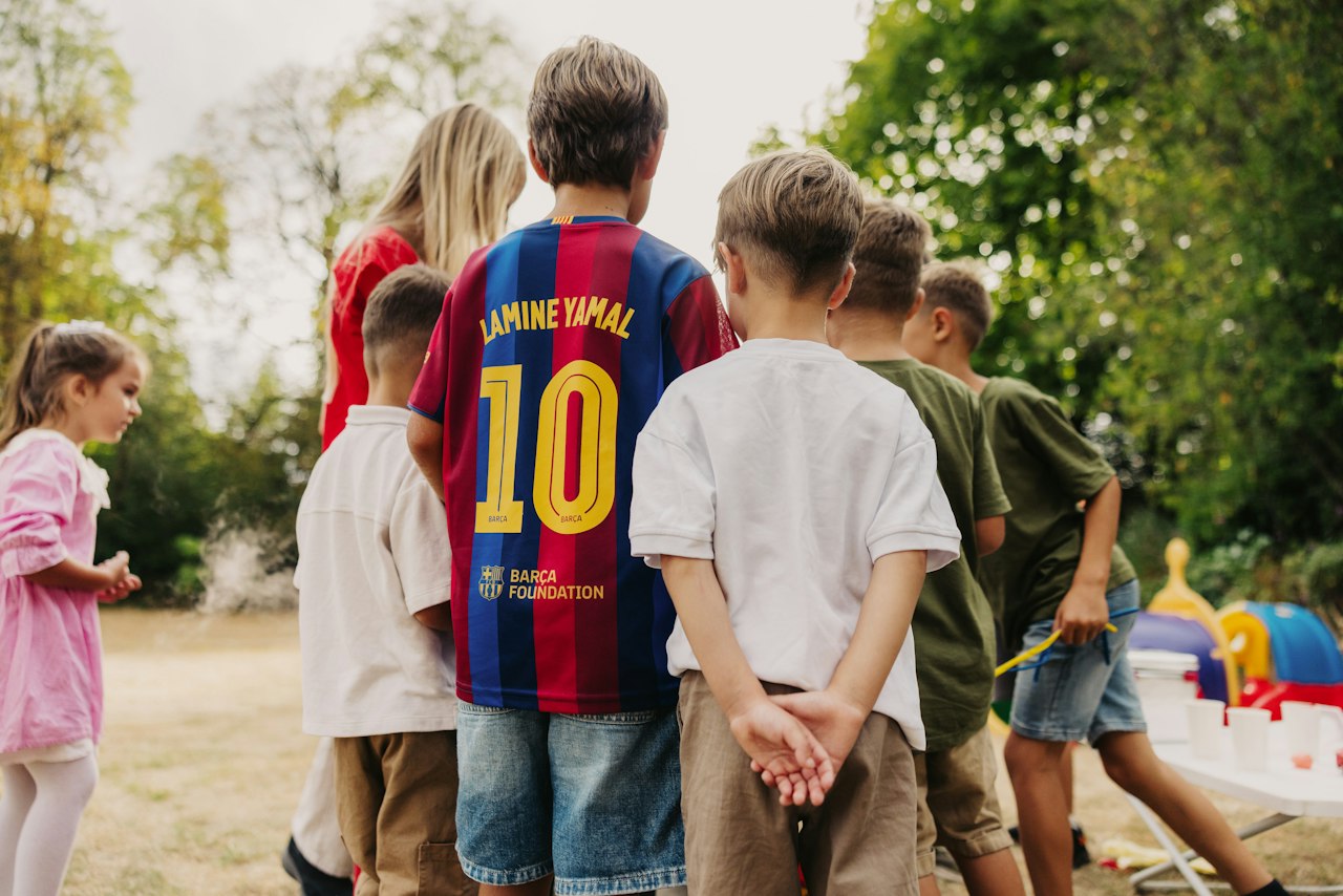a group of children playing outside, shot from behind. they gather together to peer over one boys shoulder and see what he is holding out to show them