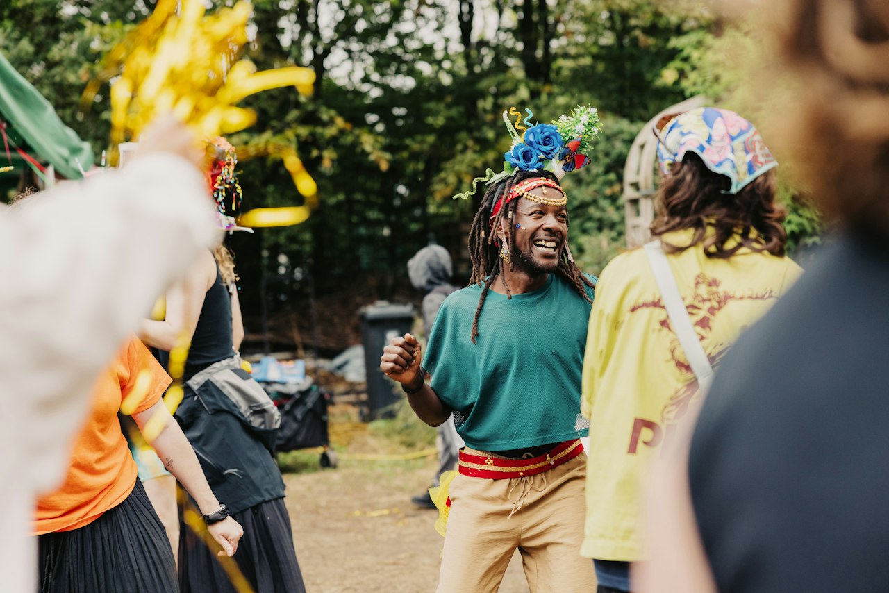A group of people dancing outside as part of a Tottenham Families Fight Back Carnival event. A person in the centre wearing a green top and a homemade headdress is smiling as golden streamers are thrown in the air.