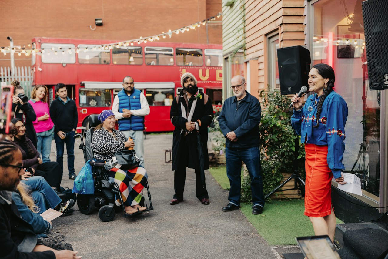 An outdoor event at which a group of people have gathered to watch someone speak. Above them is a row of glistening fairy lights and behind them a bright red double-decker bus.