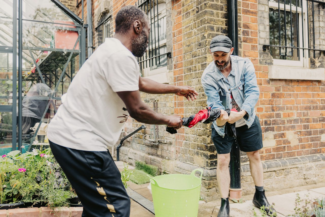 Outside in a community garden, two people are working together to wring out a black and red cloth. Towards the ground in front of the greenhouse on their left, lies a bed of green and pink flowers that have been freshly watered.