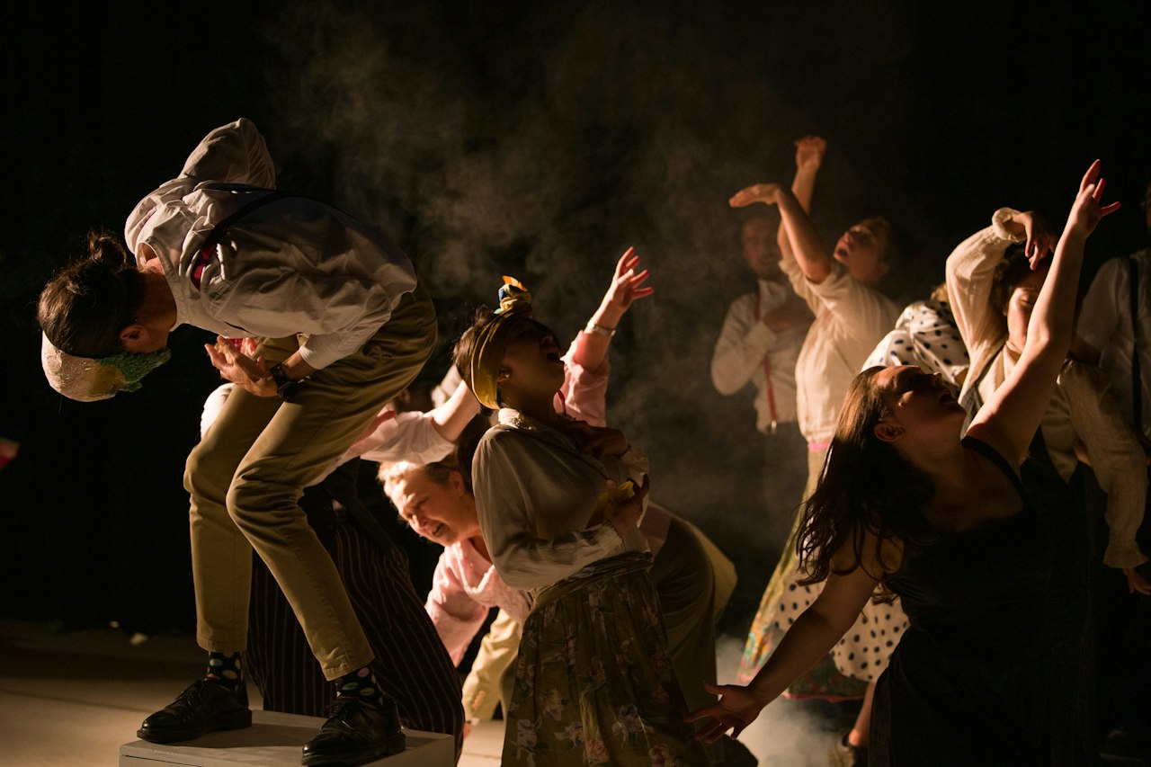 a group of people dancing on stage. the stage has low lighting and lots of dry mist, and the dancers crouch and spin low to the ground in white outfits.