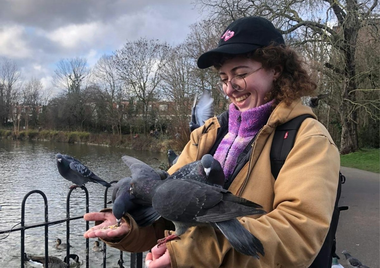a photograph of a woman with curly hair in waterproof clothing smiling while pigeons sit on her arms