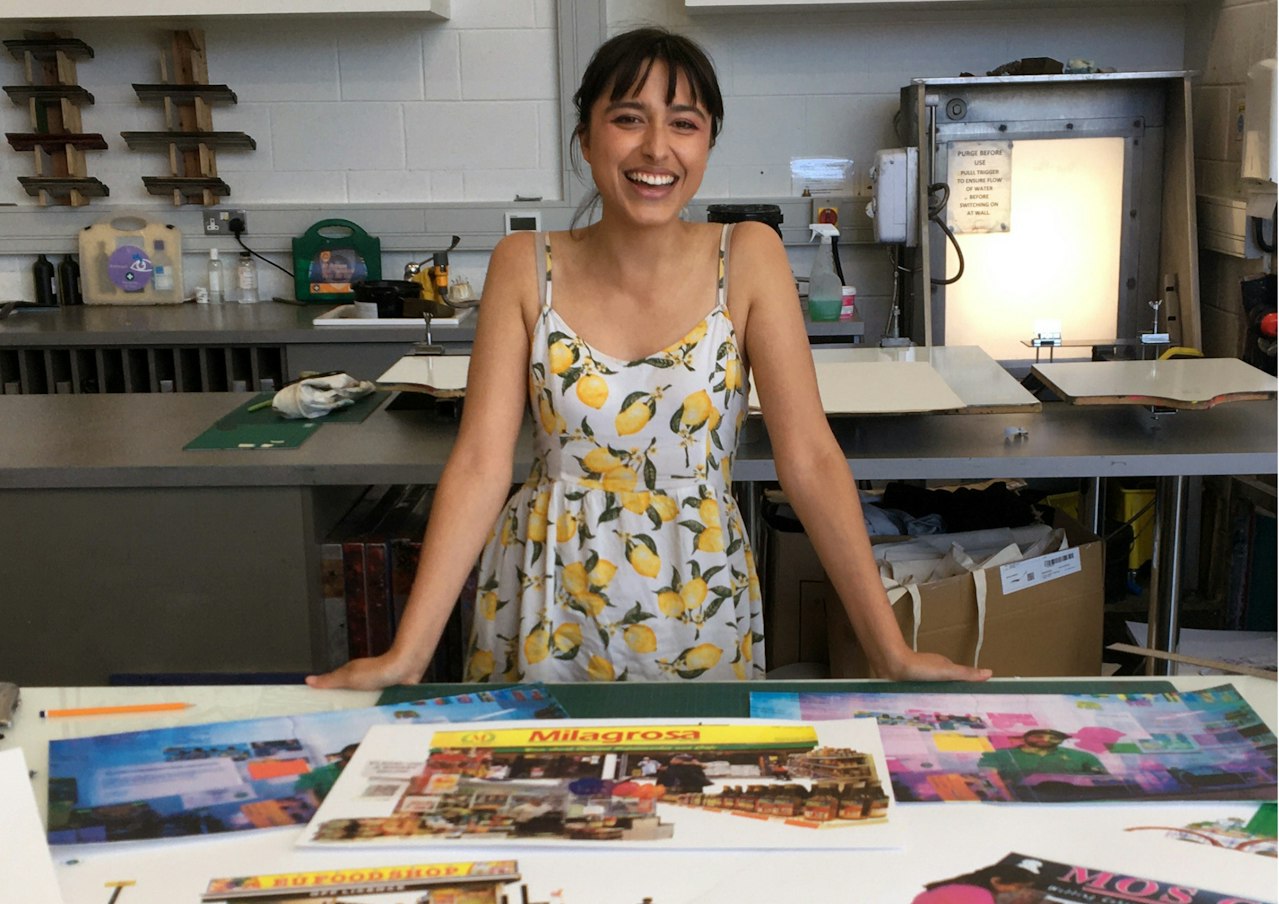 a photograph of the artist in their studio smiling in front of a table covered in photographs. She is a brunette woman in a white dress with a lemon-print on it.