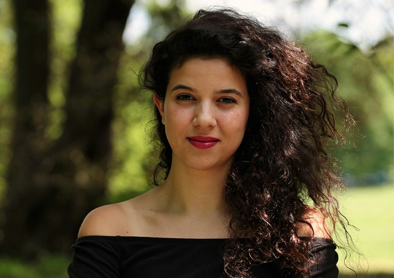 A young woman with voluminous curly brown hair and brown eyes smiles kindly into the camera, the background is a bokeh effect but you can tell she is stood outside in the sun somewhere where there are lots of trees.