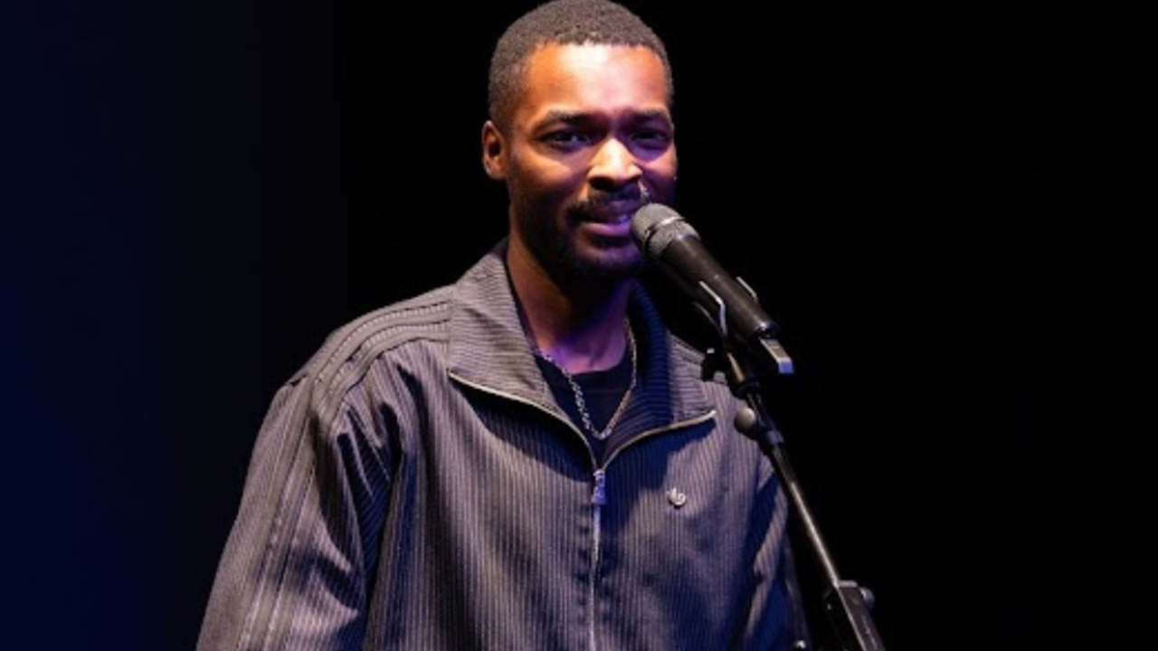 Haringey based poet, Kieron Rennie. He is a young Black man with short hair, wearing a pinstriped-black tracksuit. He is performing on stage, speaking into a microphone. There are black curtains behind him, and he is under a spotlight.