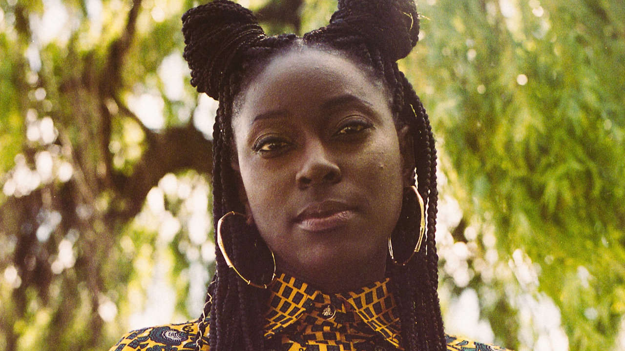 A portrait-style photograph of a young Black woman. She wears a colourful yellow and black printed shirt and has her hair in two braided buns ion top of her head. She is stood outside on a sunny day.
