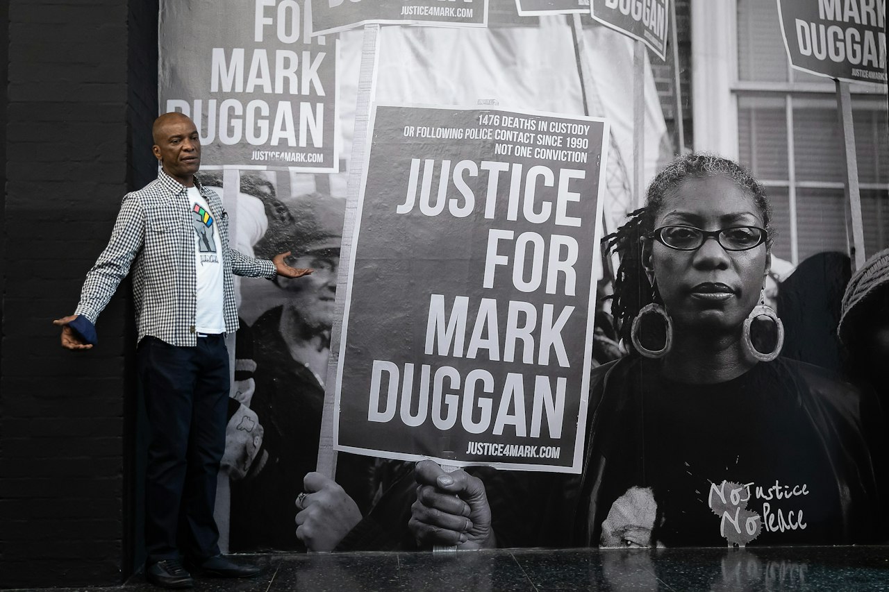 A photo of Stafford Scott, Director of Tottenham Rights, speaking on stage, in front of an archive photo which shows people holding placards reading 'Justice for Mark Duggan'.