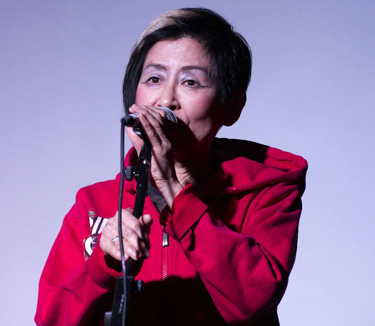 A Japanese woman with purple eyeshadow and an asymmetrical hairstyle singing into a microphone.