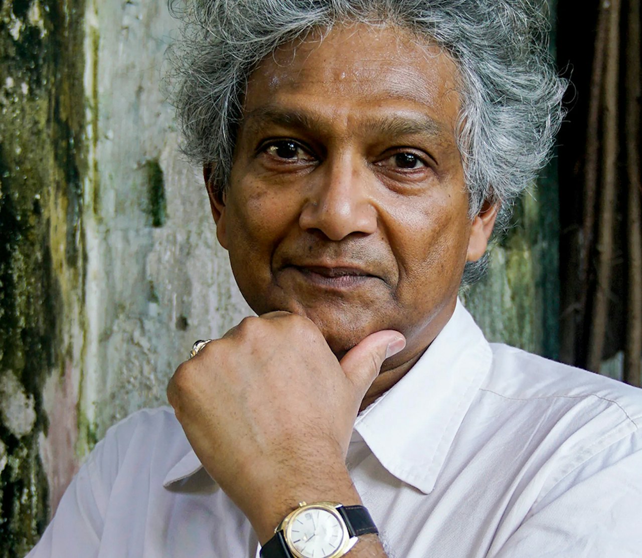 A close-up photograph of the artist Romesh, showing only his head and shoulders. He is wearing a white shirt and a watch, which you can see from his pose - one hand is raised, and his chin rests upon it as if deep in thought.