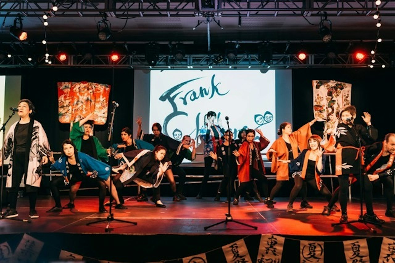 A group of people siznging and dancing together on stage. In the background can be seen a banner reading 'Frank Chickens'